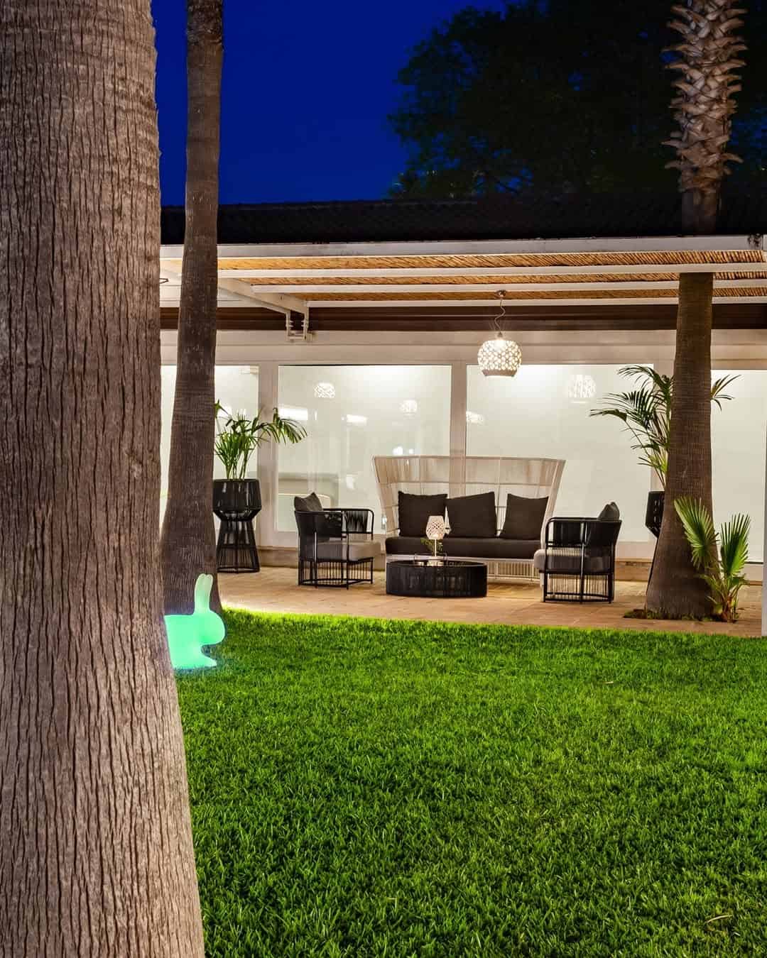 Outdoor patio at night with black wicker furniture, green lawn, and a glowing bunny-shaped light near a tree&mdash;evoking the charm of the best beach resorts in Italy for families.
