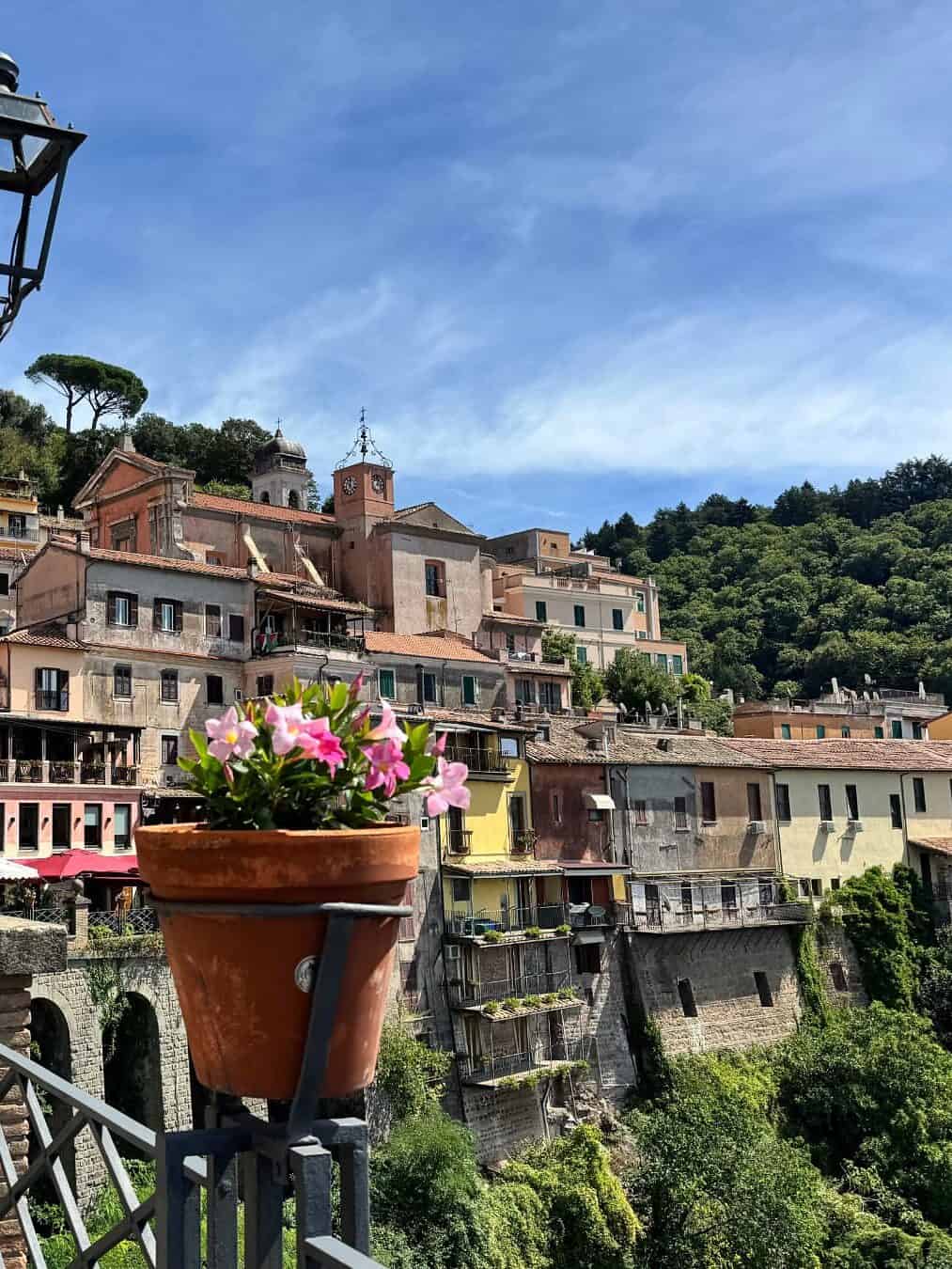 A pot of pink flowers on a railing overlooks colorful hillside buildings and lush greenery under a blue sky, capturing the charm you&rsquo;ll experience during 4 days in Castel Gandolfo.