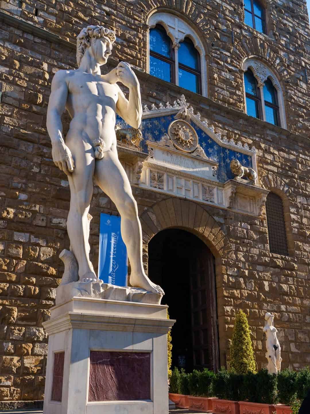 The Statue of David stands before a historic stone building with an arched doorway and ornate decorations&mdash;an iconic sight often included in the best day trips from Rome for families.