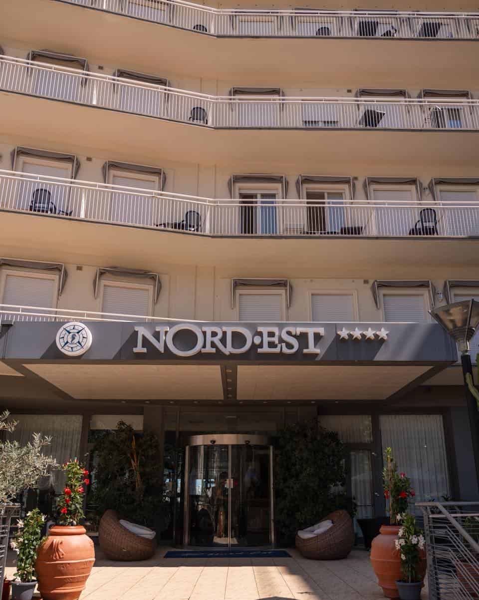 Entrance of Hotel Nord Est, one of the best beach resorts in Italy for families, with a large sign, balconies above, and potted plants by the doorway.