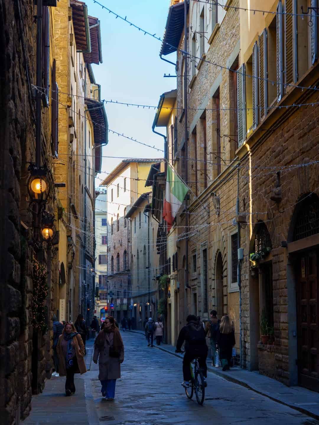 Narrow street in an old European city, with people walking, a cyclist, and buildings with hanging lights and a flag&mdash;just the kind of hidden gem you might discover on one of the best day trips from Rome for families.