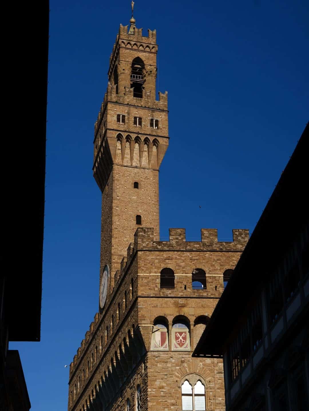 Tall stone clock tower of Palazzo Vecchio in Florence against a clear blue sky&mdash;a must-see landmark and one of the best day trips from Rome for families seeking culture and history.