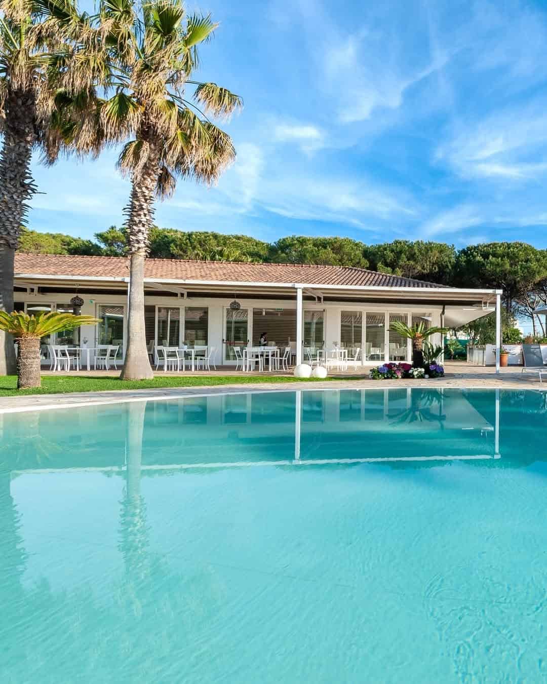 A modern house with a patio and white chairs beside a clear blue swimming pool and palm trees, evoking the relaxed luxury found at the best beach resorts in Italy for families.