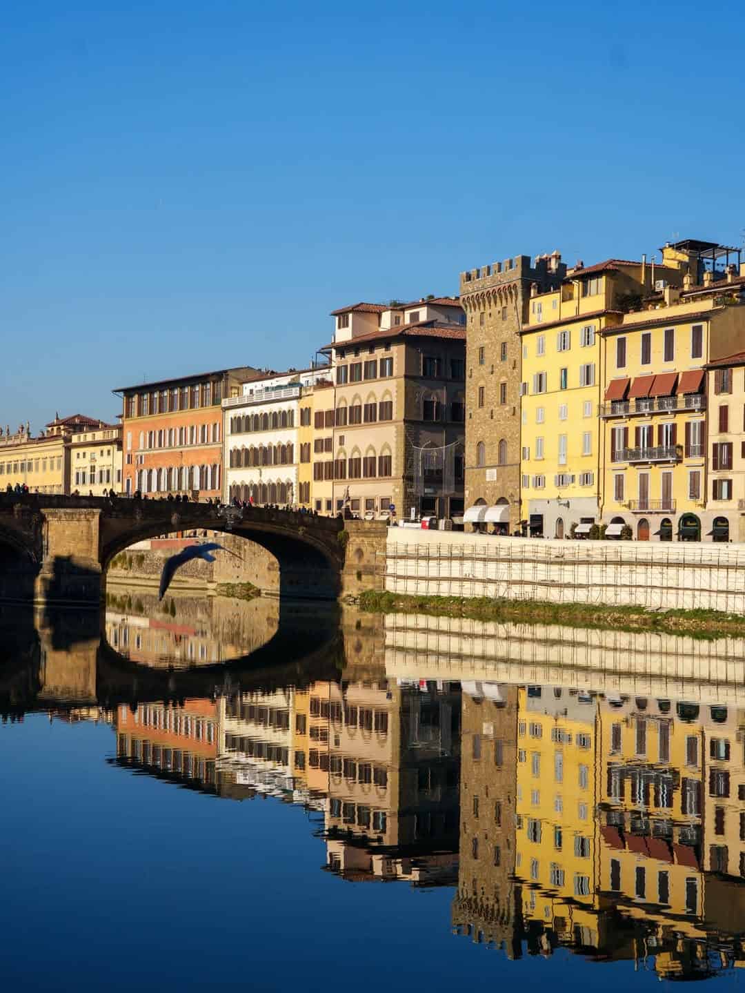 Colorful riverside buildings and a stone bridge are reflected in the calm water under a clear blue sky&mdash;an ideal spot among the best day trips from Rome for families.
