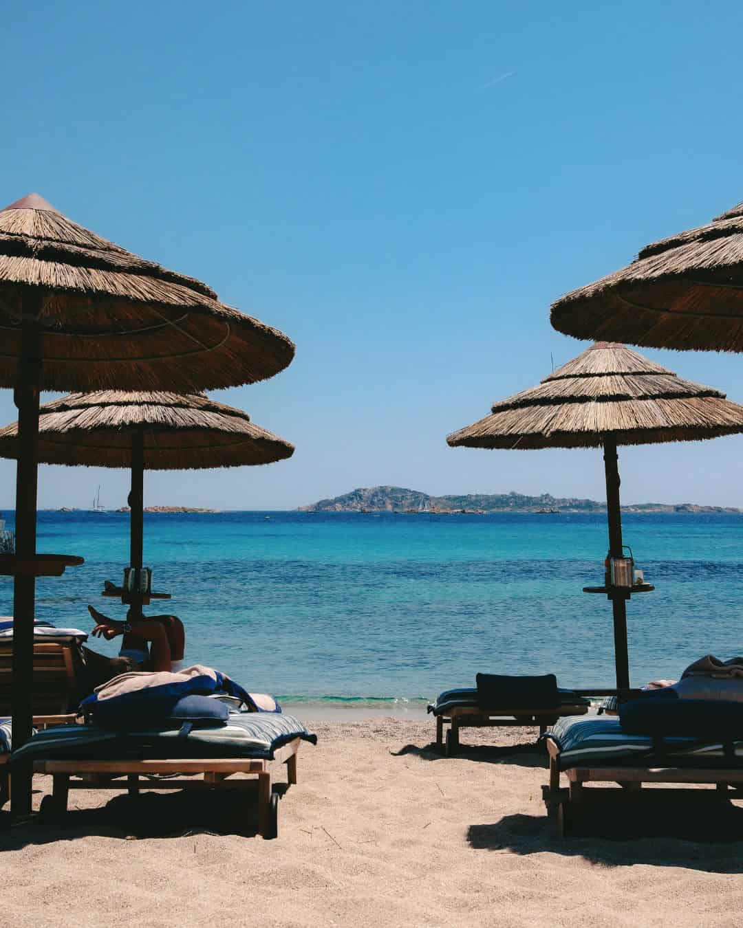 Straw beach umbrellas and lounge chairs on a sandy shore facing the calm blue sea and distant island&mdash;perfect for families seeking the best beach resorts in Italy under a clear sky.