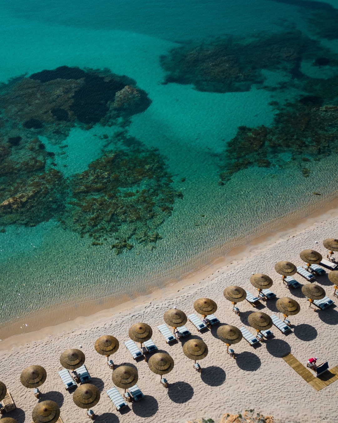 Aerial view of a sandy beach with straw umbrellas and lounge chairs, offering clear turquoise water with rocks&mdash;perfect for those seeking the best beach resorts in Italy for families.