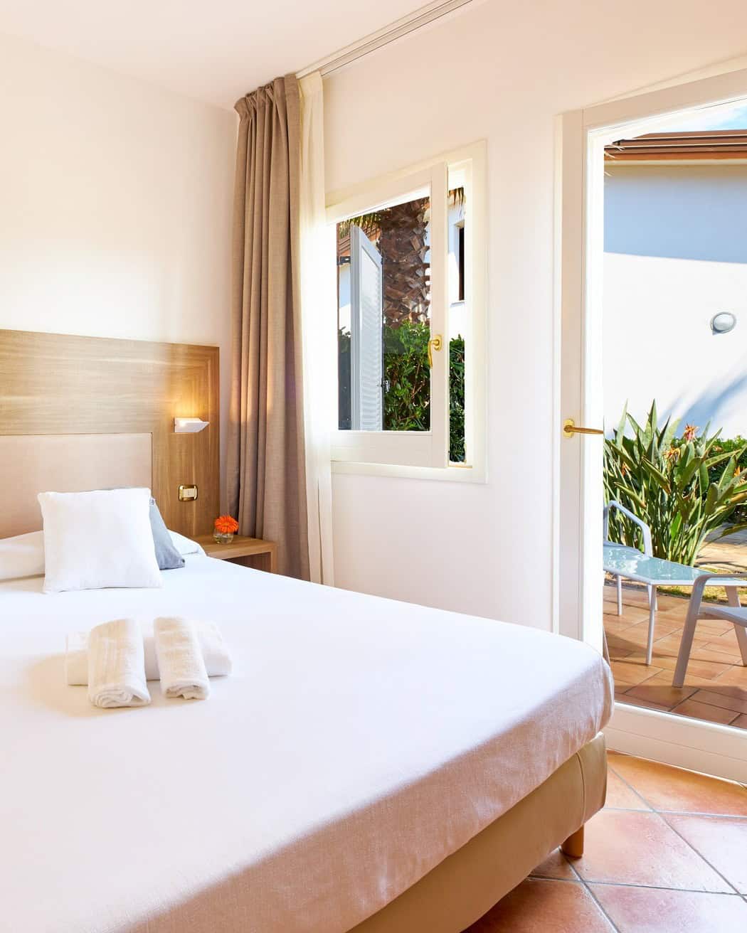 Bright bedroom with a neatly made bed, towels on top, and a door opening to a sunny outdoor patio with plants&mdash;perfect for relaxing after exploring the best beach resorts in Italy for families.