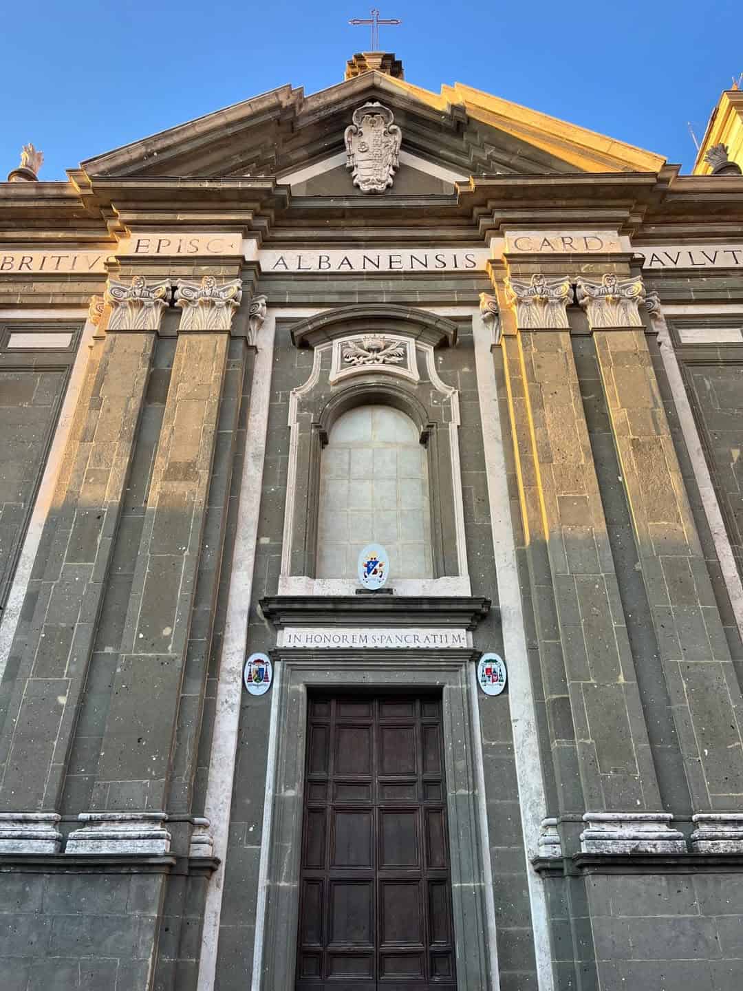 Facade of a historic stone church with tall columns, ornate details, and a large wooden door at sunset—perfect for capturing the charm you'll experience during 4 days in Castel Gandolfo.