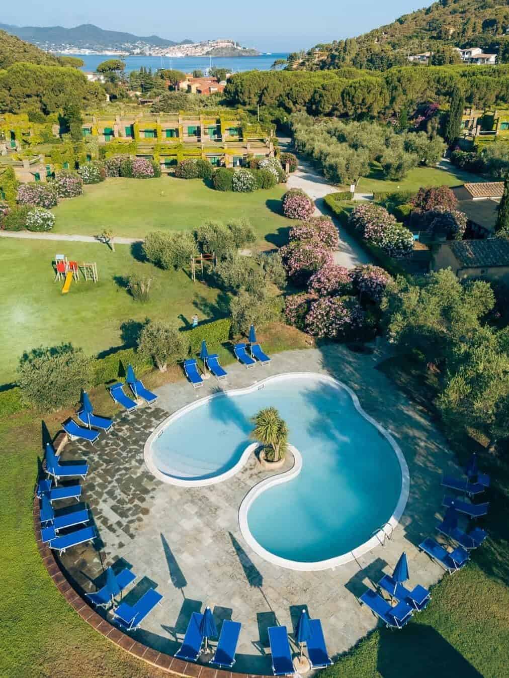 Aerial view of one of the best beach resorts in Italy for families, featuring a heart-shaped pool, blue lounge chairs, lush gardens, and distant hills.