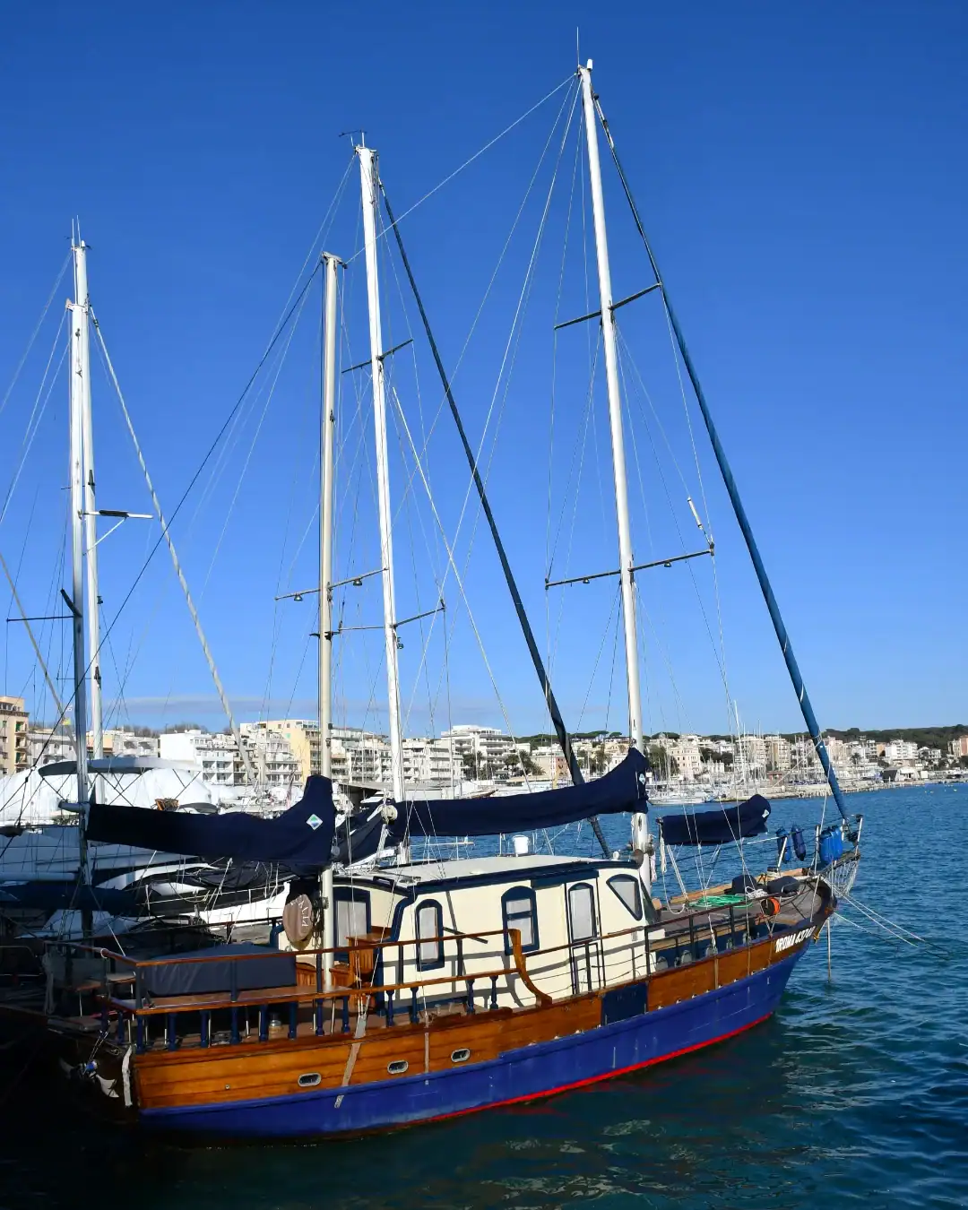 A wooden sailboat with tall masts docked in a marina, with buildings in the background under a blue sky&mdash;perfect for those seeking the best day trips from Rome for families.