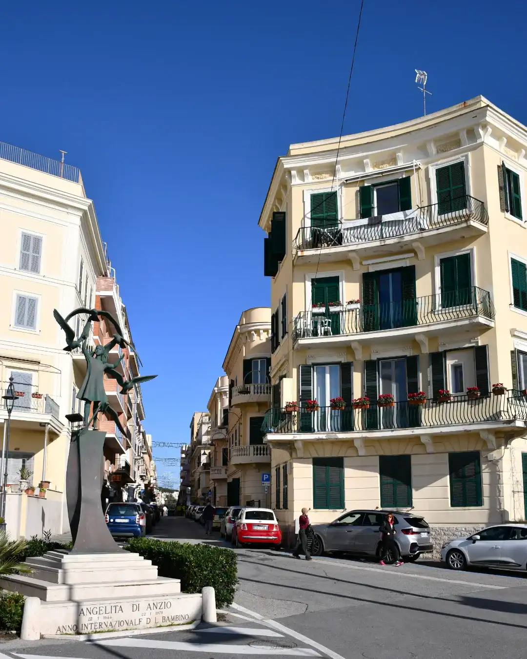 A sunny street in Anzio, Italy, with cars, people, a statue, and yellow buildings with green shutters&mdash;perfect for one of the best day trips from Rome for families.