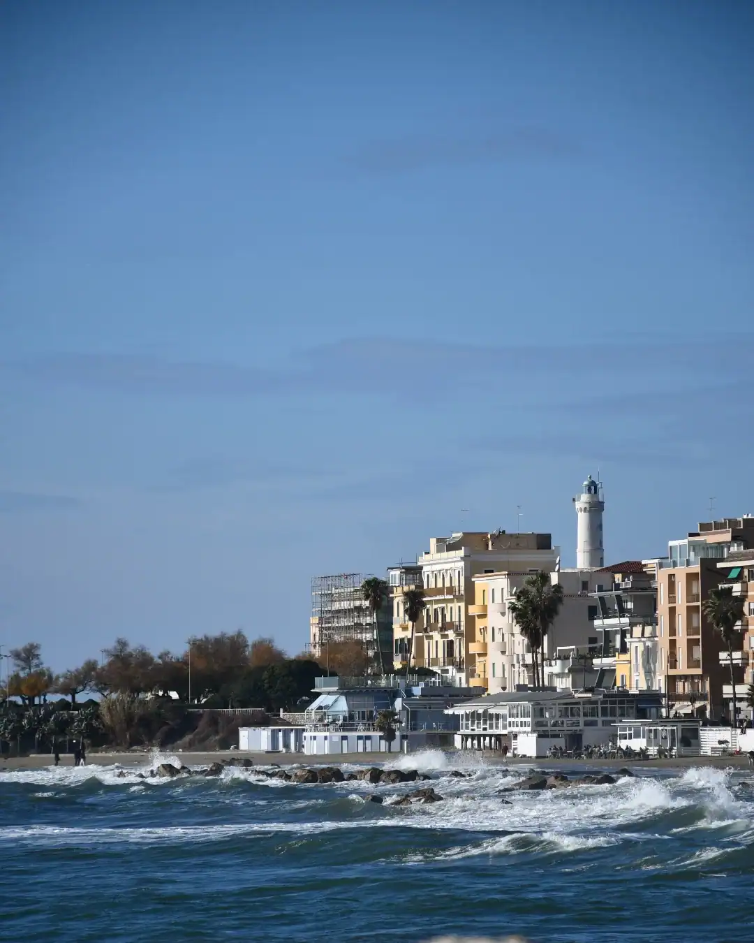 Coastal buildings and a lighthouse by the sea under a clear blue sky with waves in the foreground&mdash;an ideal spot reminiscent of the best day trips from Rome for families seeking seaside adventures.