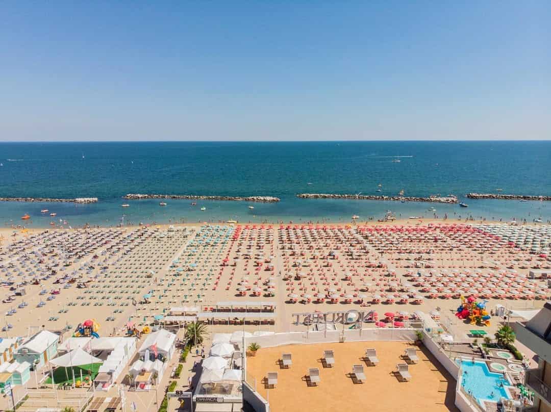 Aerial view of a crowded beach with rows of colorful umbrellas and a clear blue sea under a sunny sky, capturing the lively spirit often found at the best beach resorts in Italy for families.