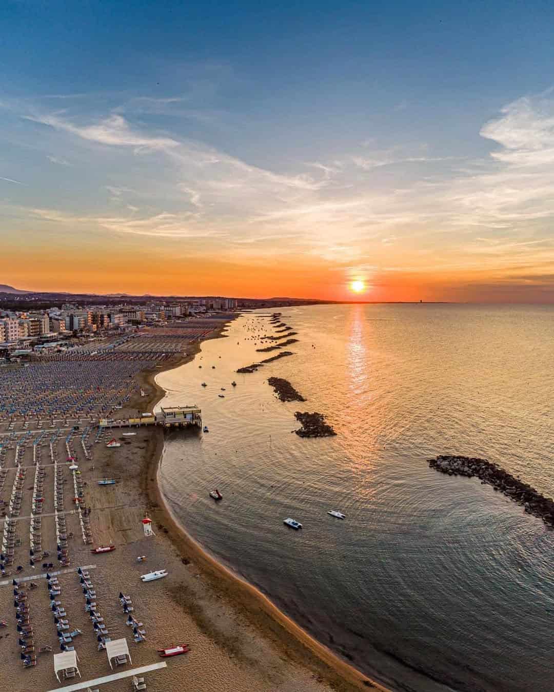 Aerial view of a beach with rows of umbrellas at sunset, city buildings and calm sea in the background&mdash;perfect for those seeking the best beach resorts in Italy for families.