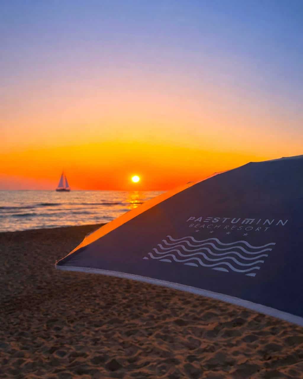 A sailboat glides on the sea at sunset, with a beach umbrella labeled "PAESTUMINN BEACH RESORT" in the foreground&mdash;one of the best beach resorts in Italy for families.