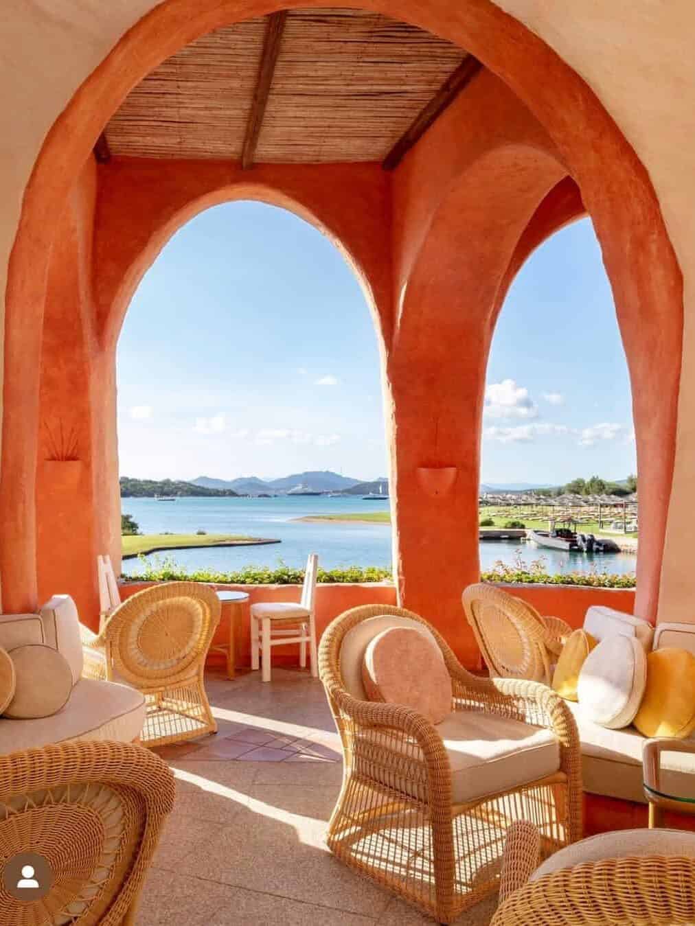 Sunny terrace with wicker chairs, coral arches, and a scenic view of water and distant hills&mdash;perfect for relaxing family moments at one of the best beach resorts in Italy for families.
