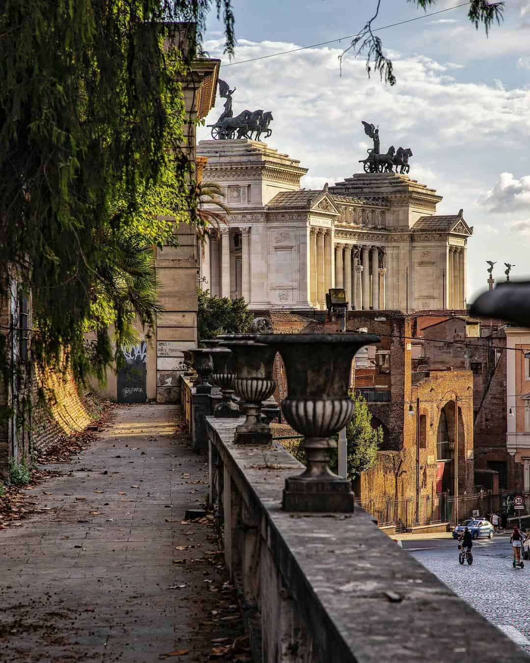 A stone walkway overlooks historic Roman buildings and statues under a partly cloudy sky, making it one of the best day trips from Rome for families seeking culture and scenic views.