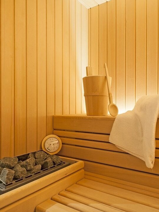 Wooden sauna interior with towel, bucket, ladle, sauna stones, and a thermometer on a bench&mdash;offering relaxation reminiscent of the best beach resorts in Italy for families.