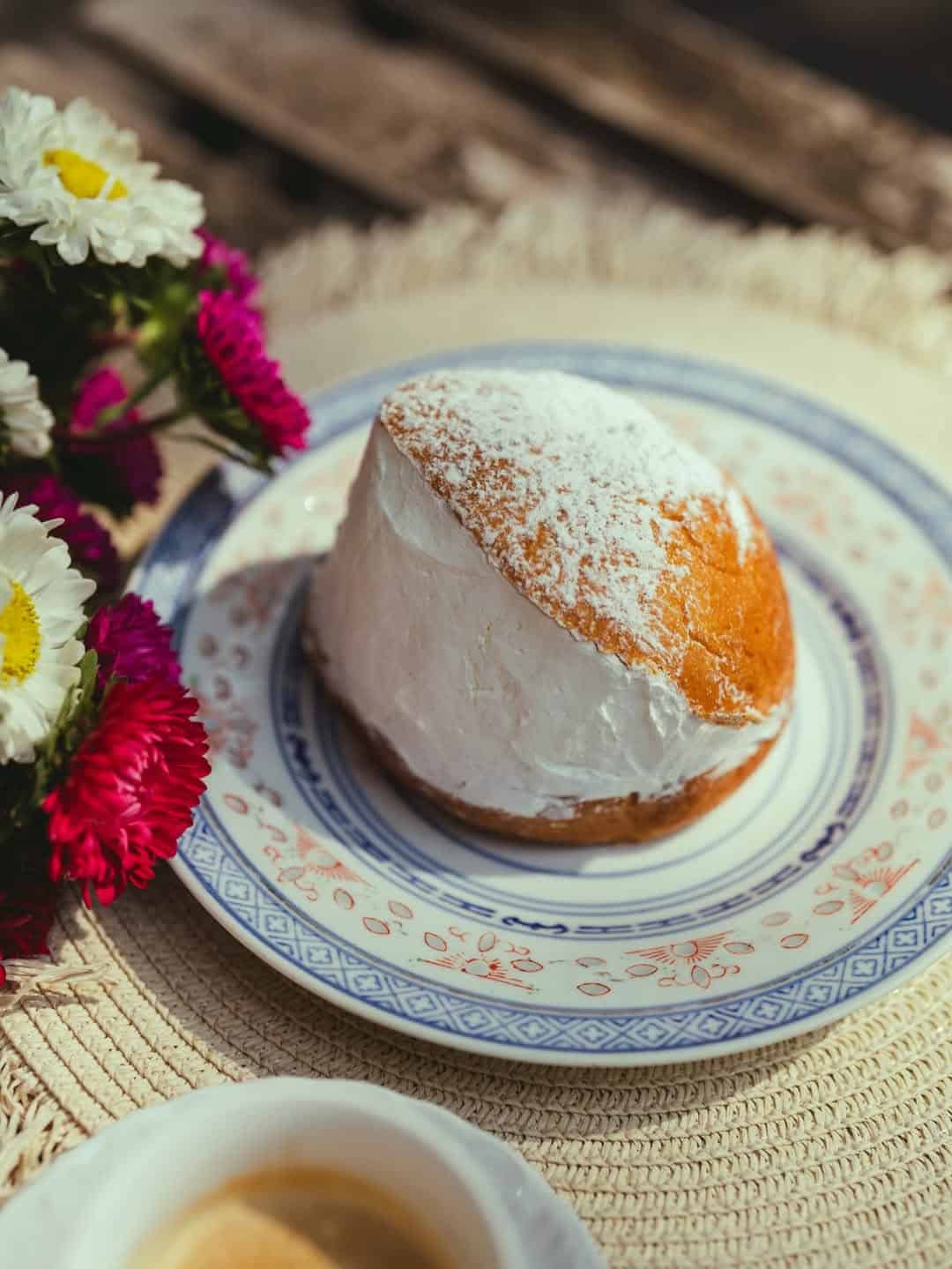 A cream-filled pastry dusted with powdered sugar on a decorative plate, beside colorful flowers—perfect inspiration for those seeking treats at Gluten Free Restaurants in Rome.