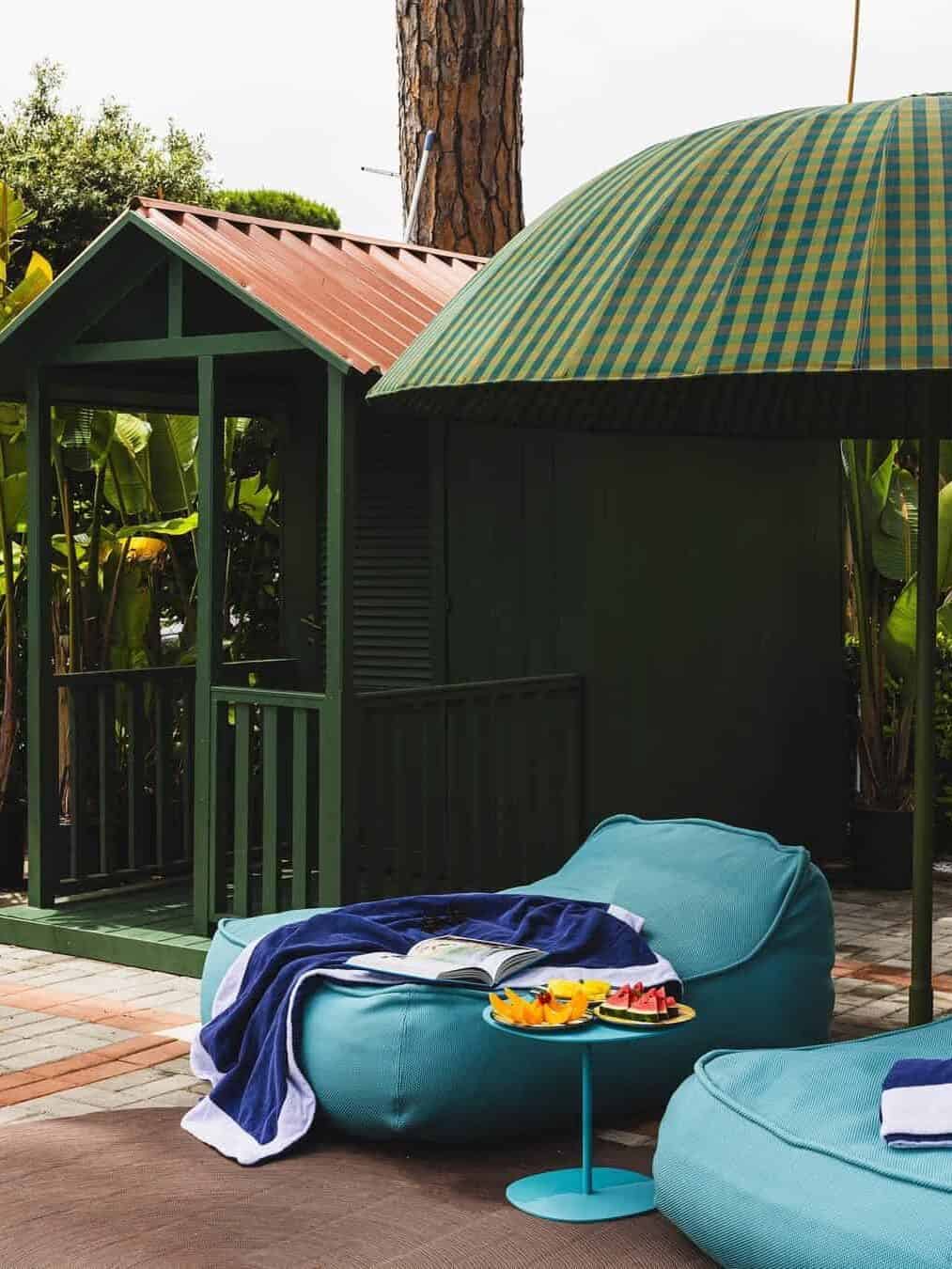 Two turquoise outdoor loungers with a towel, book, and snacks beside a green shed and large umbrella evoke the relaxing vibe of the best beach resorts in Italy for families.