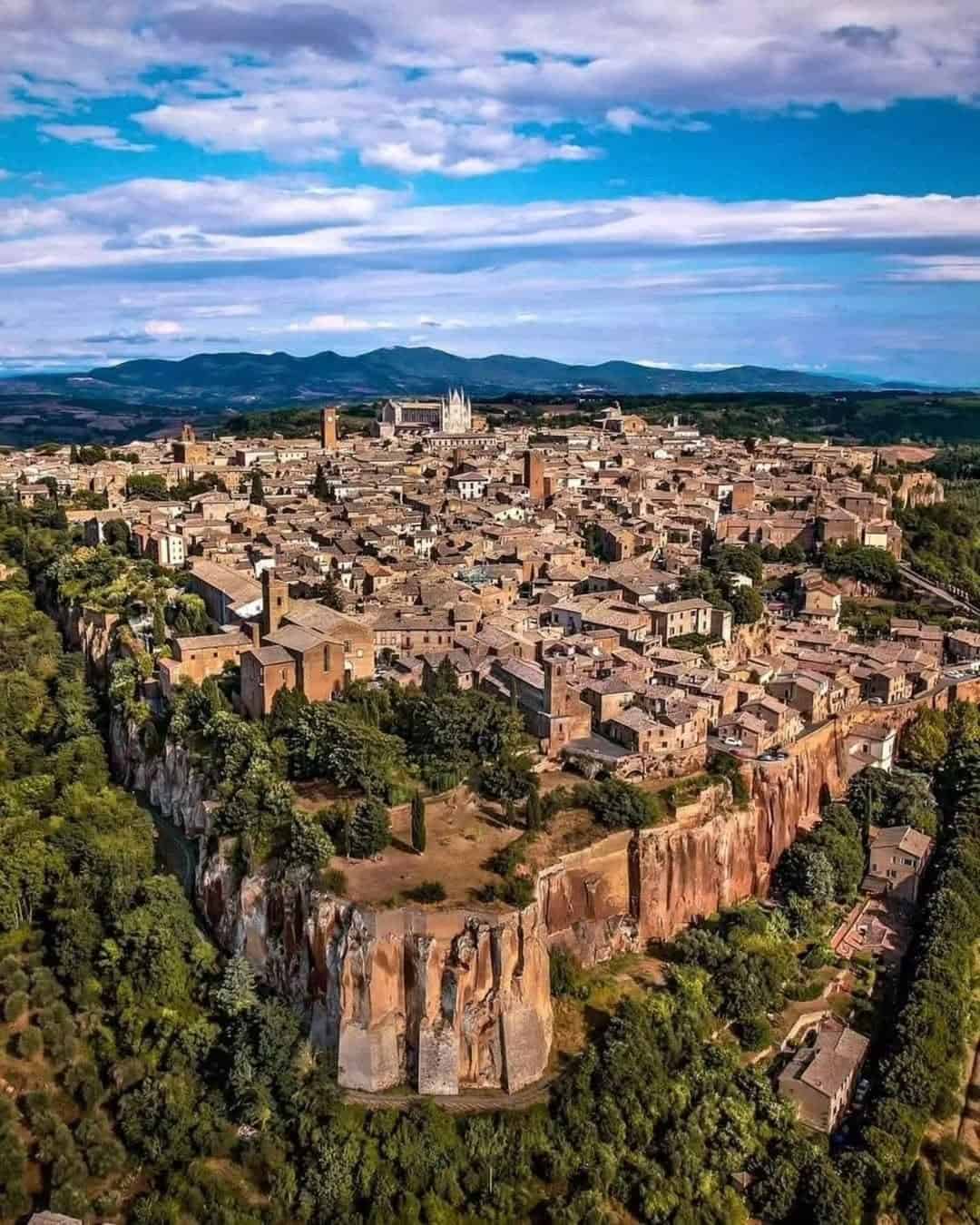 Aerial view of a historic Italian hilltop town surrounded by cliffs, trees, and distant mountains&mdash;one of the best day trips from Rome for families seeking scenic beauty and adventure.