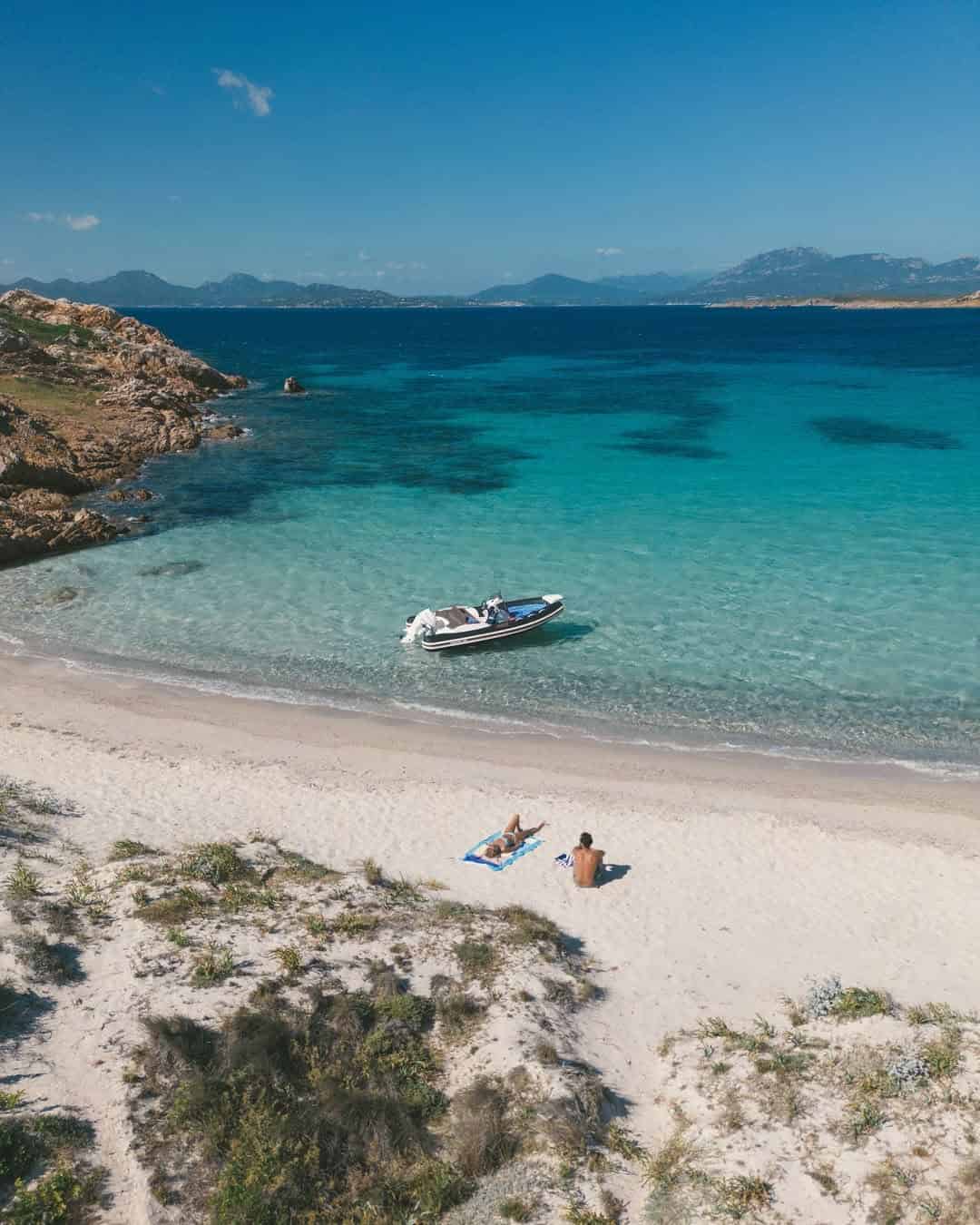 A sandy beach with two people sunbathing and a small boat anchored in the clear blue water, perfect for those seeking the best beach resorts in Italy for families.