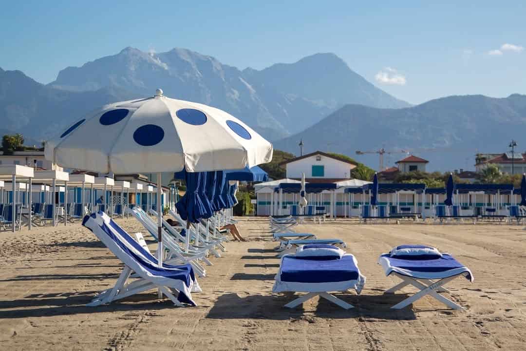 Blue and white beach chairs and umbrellas line the sandy shore, with mountains and buildings in the background&mdash;perfect scenery at one of the best beach resorts in Italy for families.