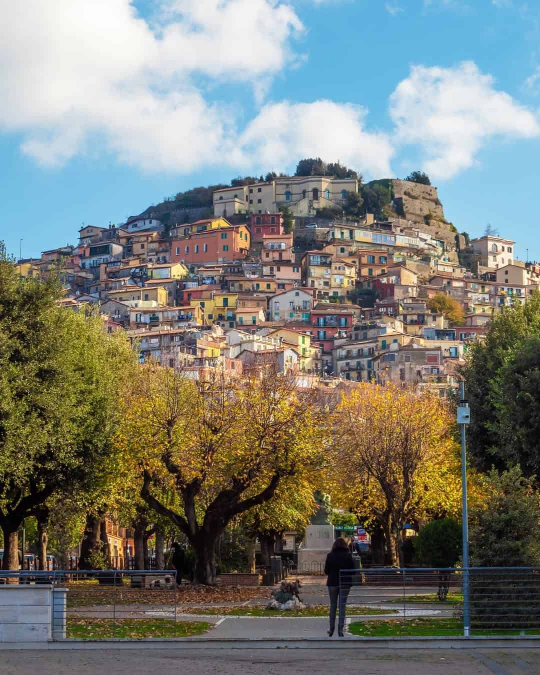 Colorful hillside houses above autumn trees and a person walking in a sunny, scenic town under a blue sky evoke the charm of spending 4 days in Castel Gandolfo.