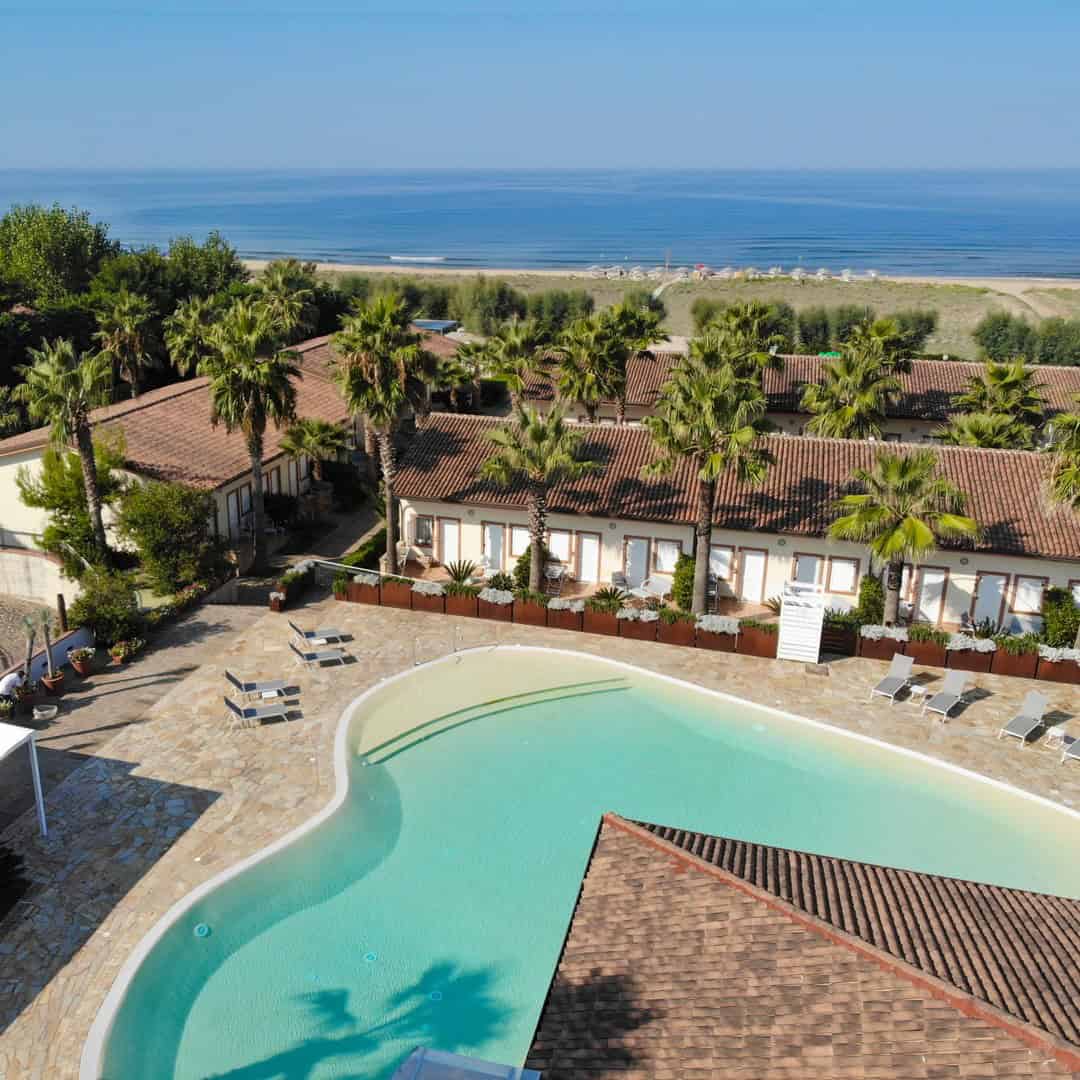 Aerial view of one of the best beach resorts in Italy for families, featuring a pool, palm trees, and villas near a sandy beach and the ocean.