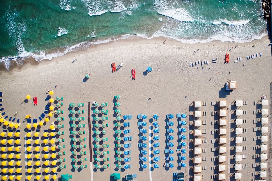 Aerial view of a beach with rows of colorful umbrellas and sunbeds by the ocean waves, capturing the lively atmosphere found at the best beach resorts in Italy for families.