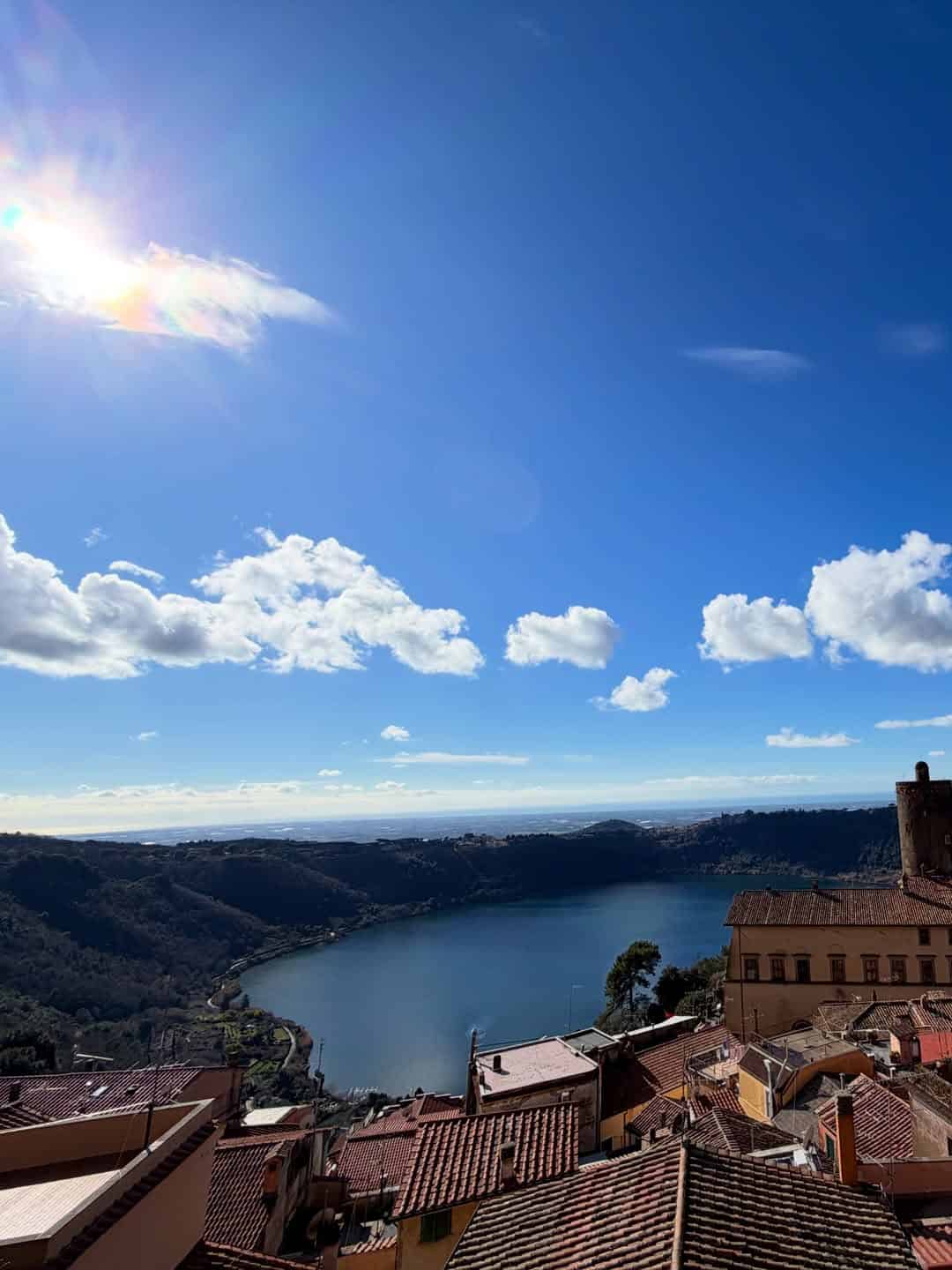 Rooftops overlook a lake and hills under a bright blue sky with scattered clouds and sunlight, capturing the serene charm you'll experience during 4 days in Castel Gandolfo.