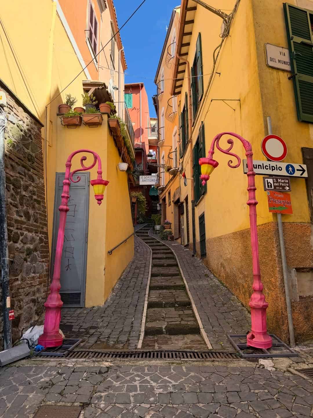 A narrow stone alley with steps, flanked by yellow buildings and two whimsical pink street lamps at the entrance—an inviting scene you might discover during 4 days in Castel Gandolfo.
