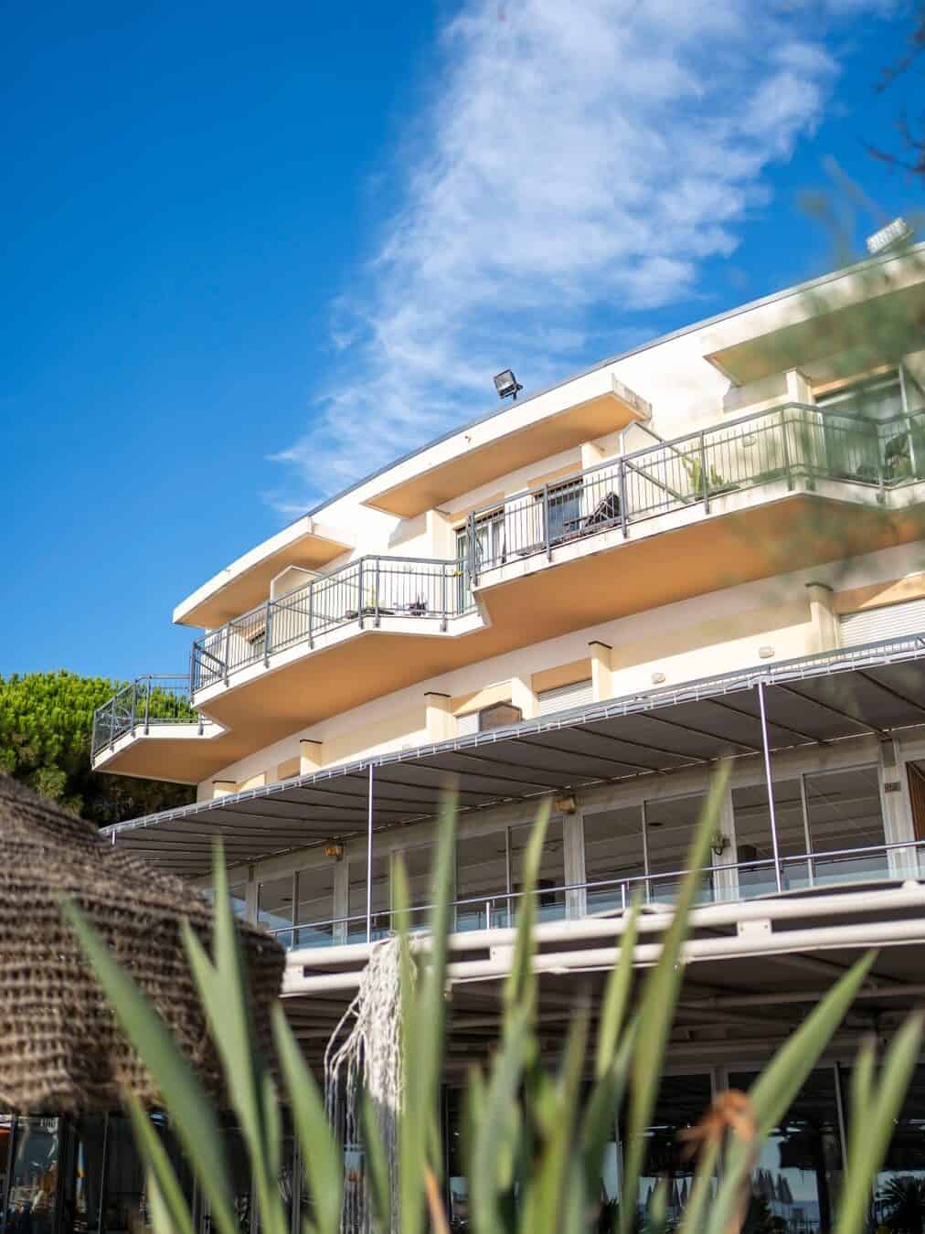 Modern building with balconies against a blue sky, framed by greenery and thatched structures in the foreground&mdash;evoking the relaxed charm of the best beach resorts in Italy for families.