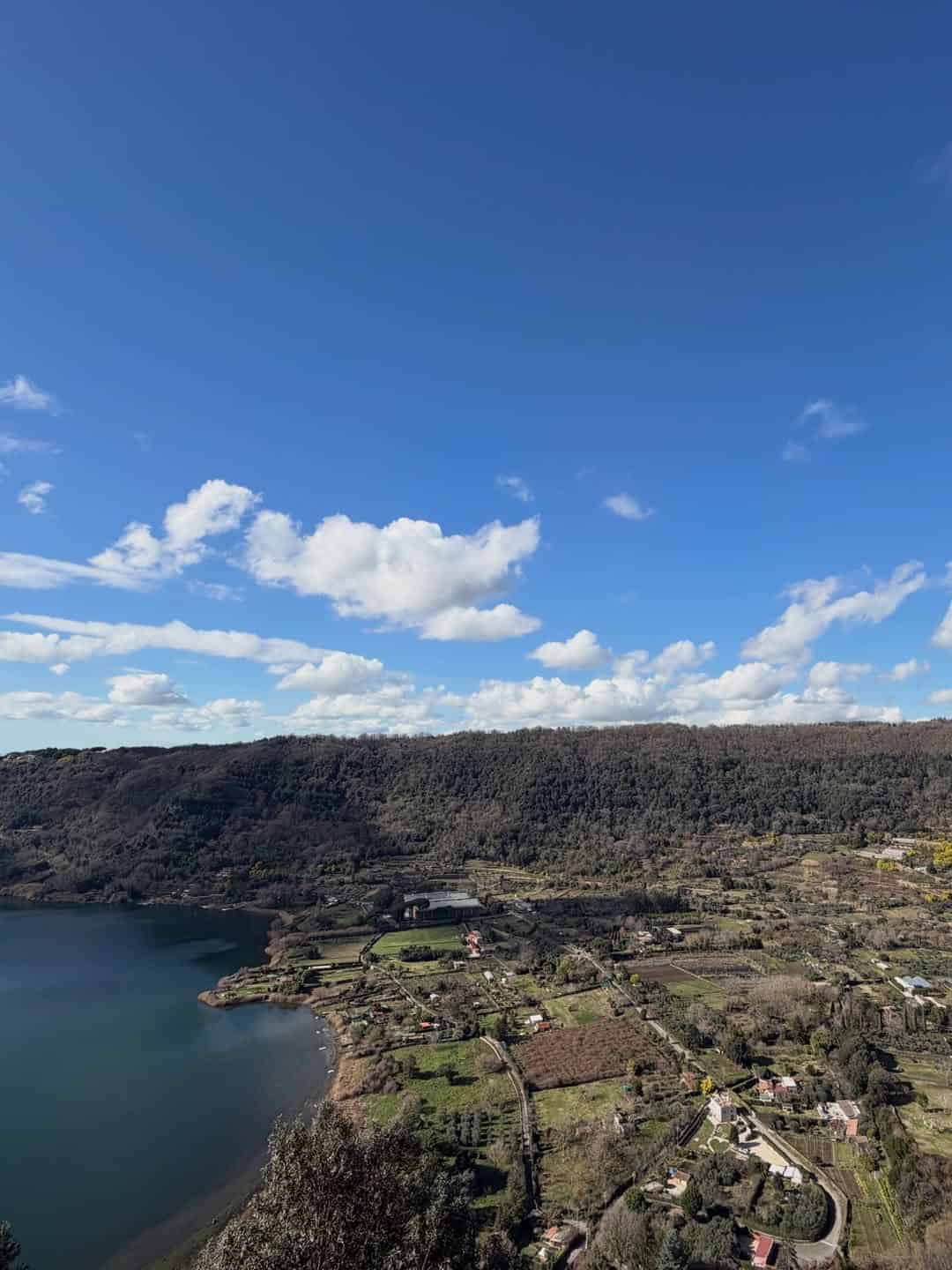 A scenic view of a lake, green fields, and scattered houses under a blue sky with fluffy clouds, perfect for spending 4 days in Castel Gandolfo.