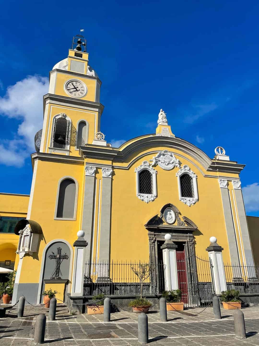 Yellow church with a clock tower against a clear blue sky; potted plants and a cross in front&mdash;an idyllic stop on one of the best day trips from Rome for families.