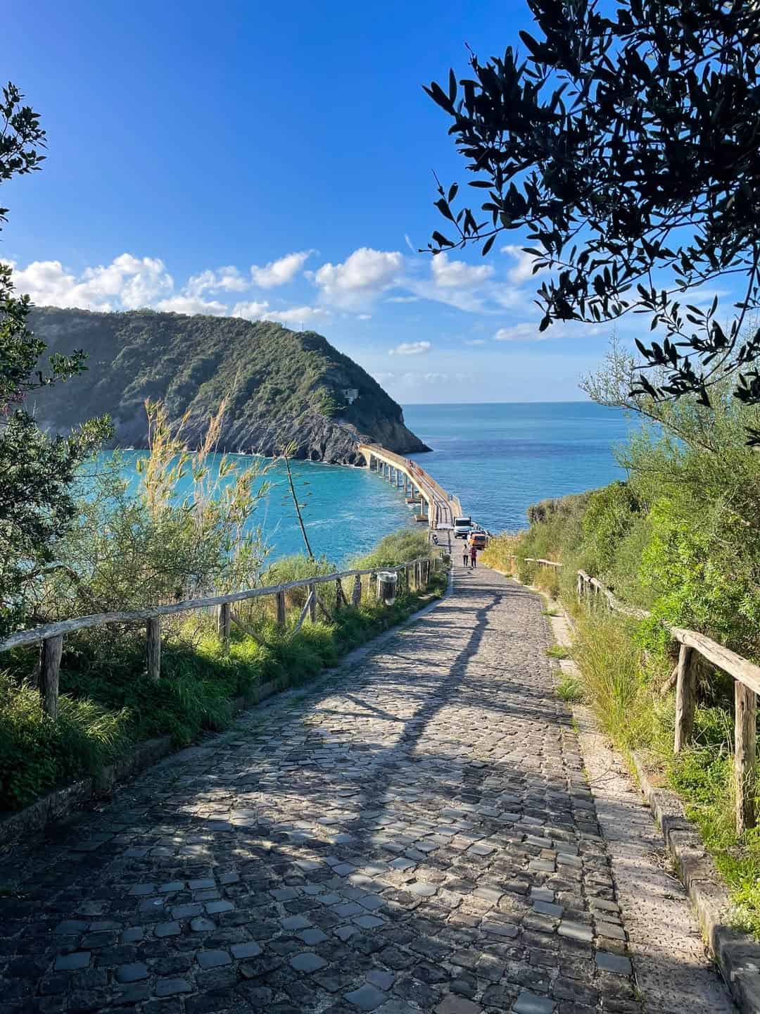 Stone path with railings leading downhill toward a curved bridge over blue sea, surrounded by green hills&mdash;an idyllic spot that's among the best day trips from Rome for families.