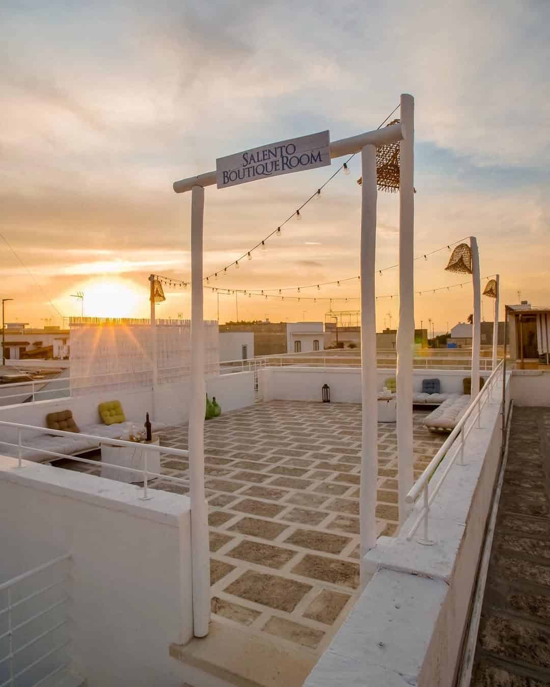 Rooftop terrace with string lights, lounge seating, and a sunset sky; sign reads "Salento Boutique Room," offering a stylish retreat inspired by the best beach resorts in Italy for families.