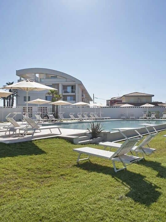 Lawn with lounge chairs, pool, umbrellas, and a modern building under a clear blue sky&mdash;perfect for those seeking the best beach resorts in Italy for families.