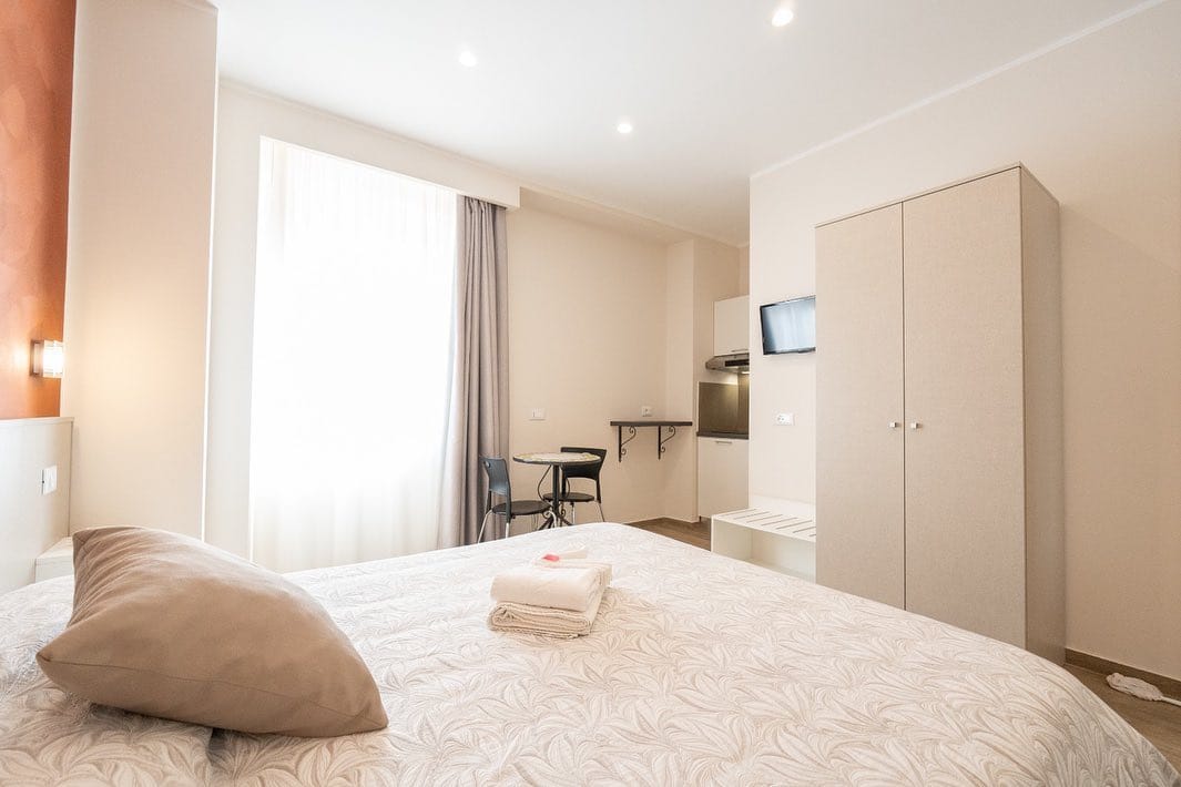 Bright, minimal hotel room with a bed, wardrobe, TV, kitchenette, and a window with sheer curtains&mdash;an inviting retreat inspired by the best beach resorts in Italy for families.