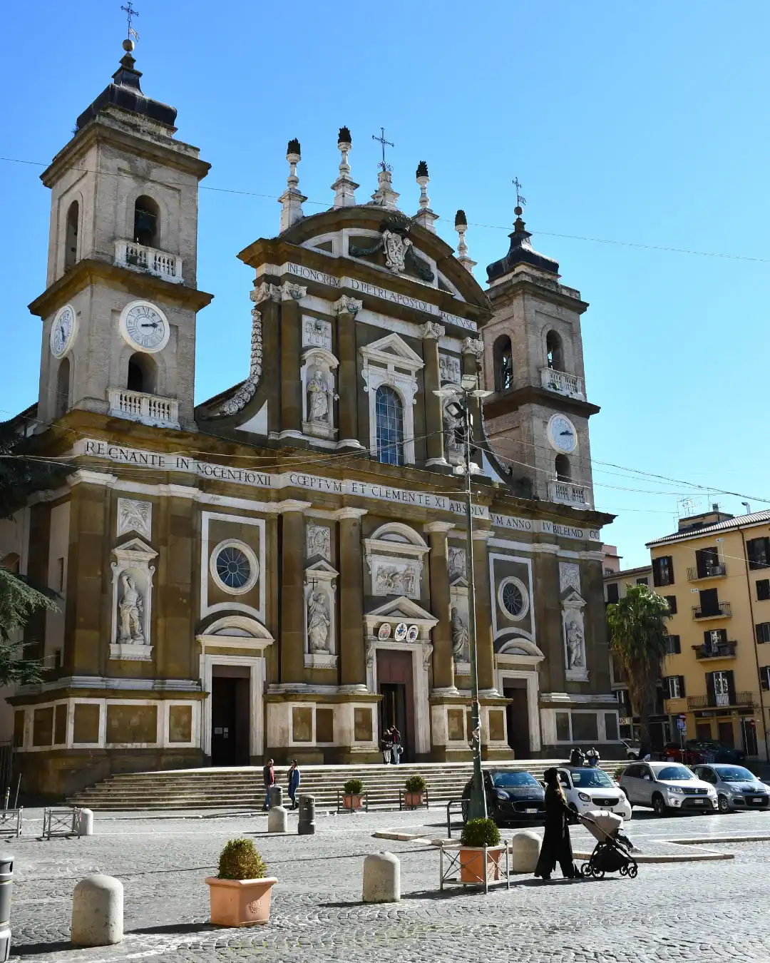 A grand church with twin towers and statues, with people walking and a stroller in the sunny plaza out front&mdash;an ideal stop on one of the best day trips from Rome for families.