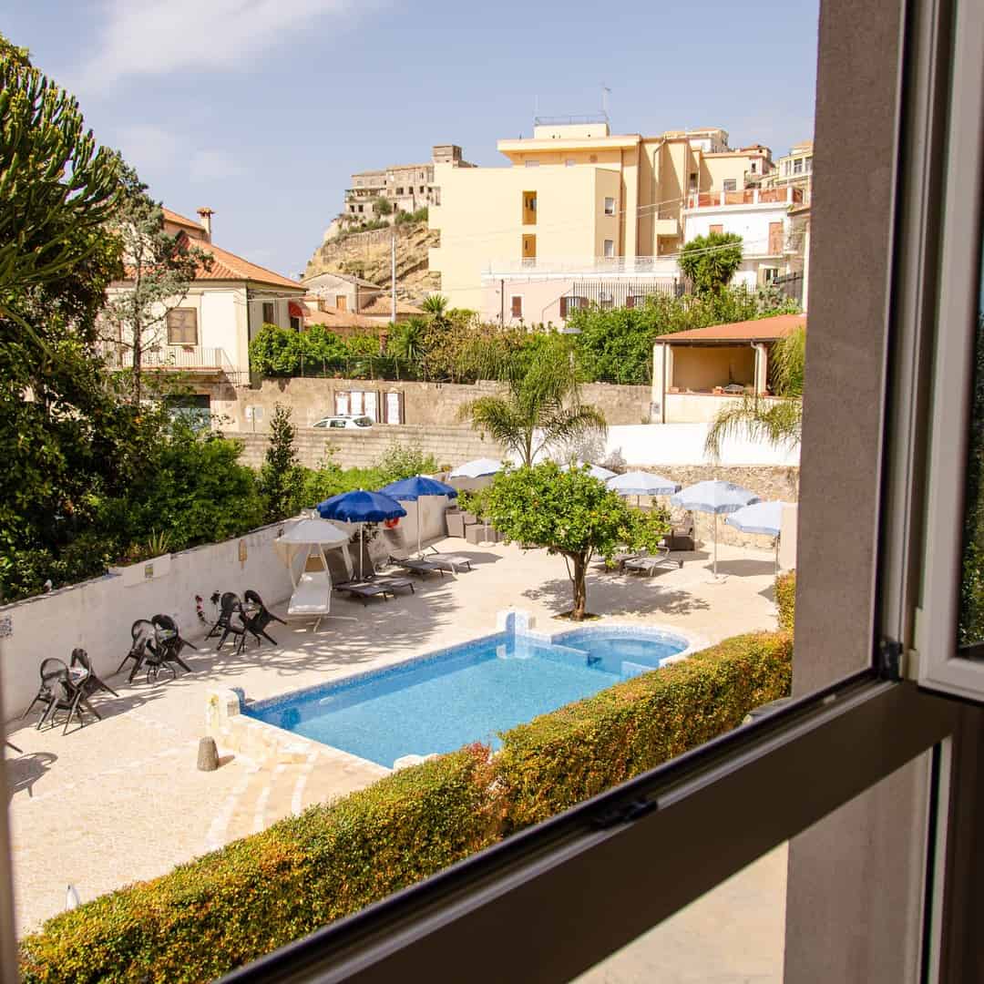 View from a window overlooking a small outdoor pool with lounge chairs and umbrellas in a sunny courtyard, reminiscent of the relaxing atmosphere found at the best beach resorts in Italy for families.