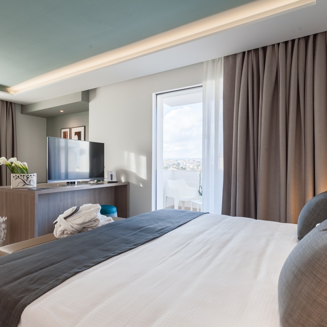 A modern hotel room with a neatly made bed, TV, and balcony with outdoor seating overlooking the city&mdash;perfect for travelers seeking comforts found in the best beach resorts in Italy for families.