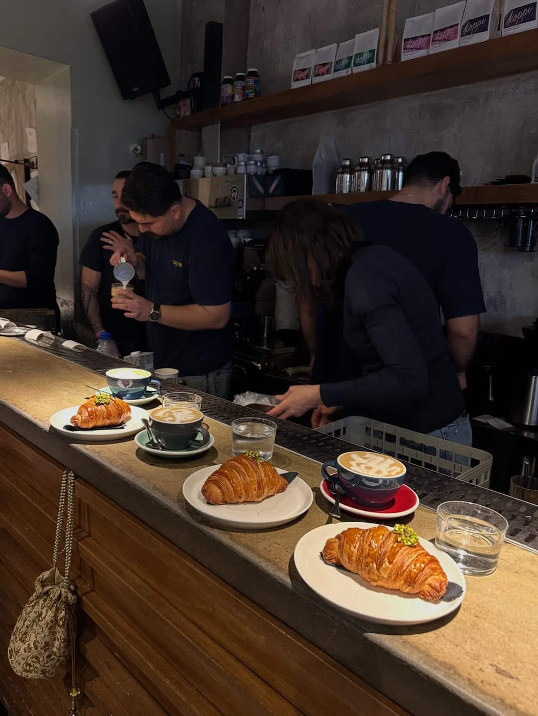 Four croissants and coffee cups on a counter, with baristas preparing drinks behind in a cozy café setting, reminiscent of the warm hospitality found in some Gluten Free Restaurants in Rome.