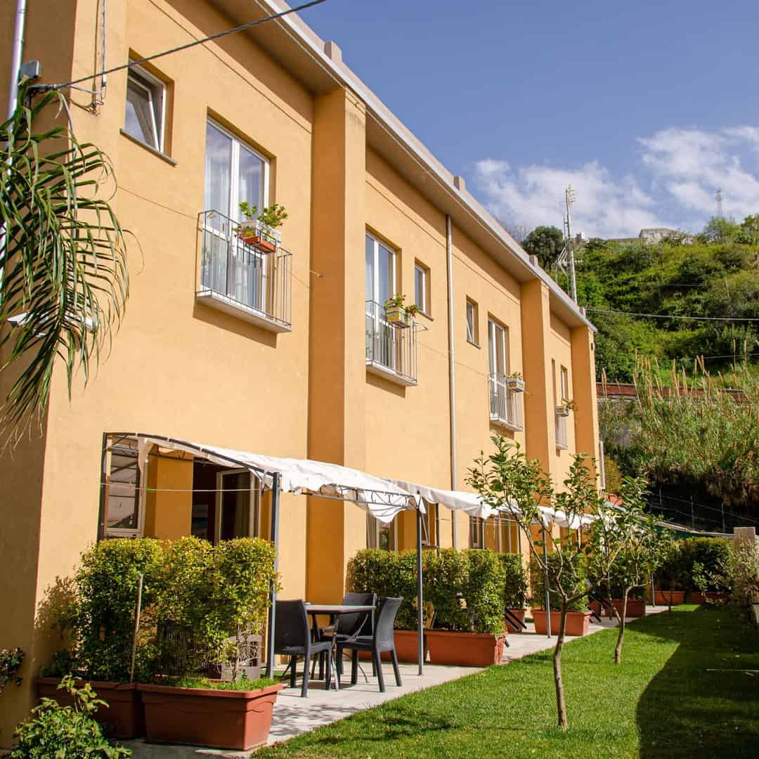Yellow apartment building with balconies, patio tables, potted plants, and green grass under a blue sky&mdash;reminiscent of the relaxed charm found at the best beach resorts in Italy for families.