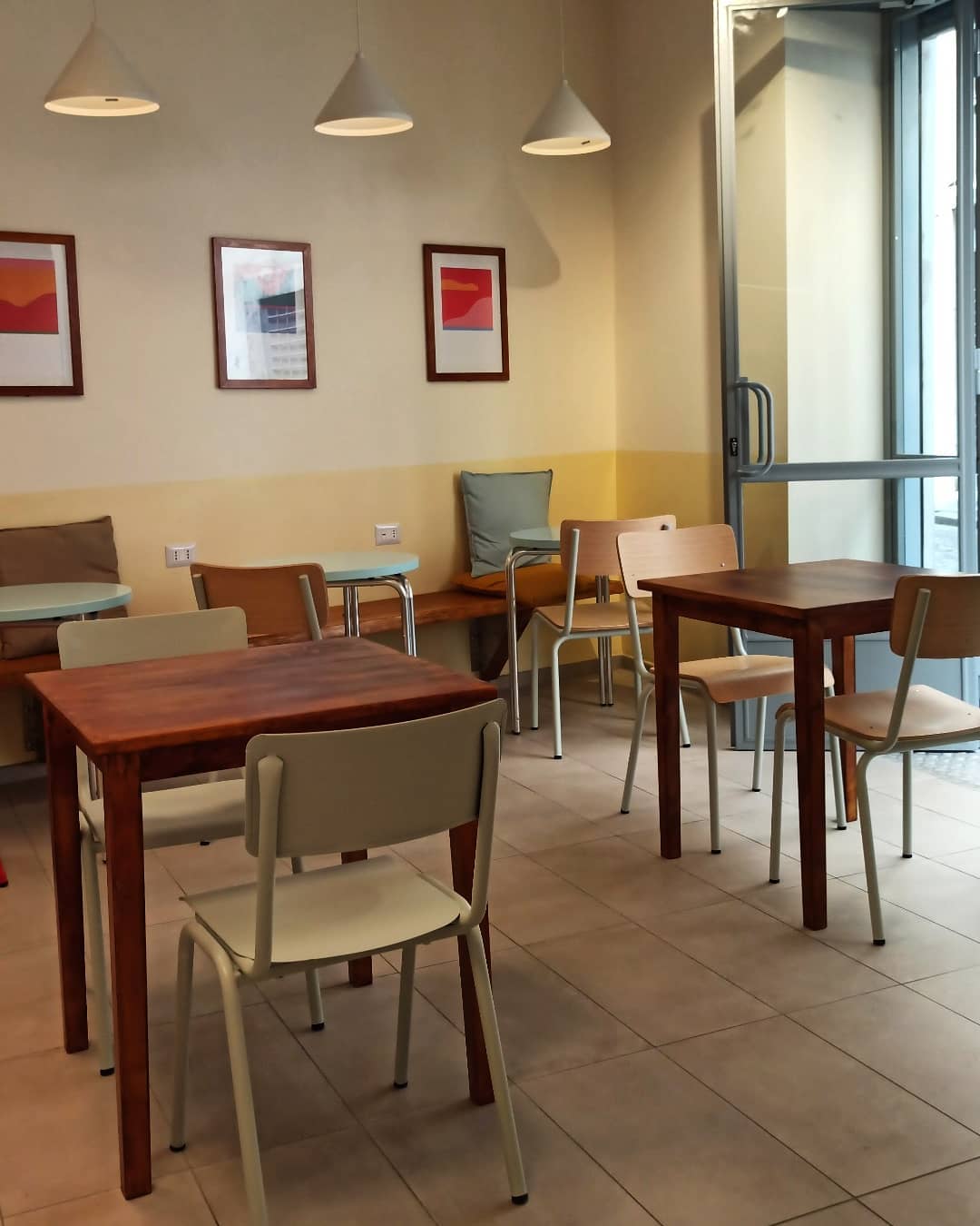 Small, modern cafe with wooden tables, light chairs, wall art, pendant lights, and a large glass door—an inviting spot among Gluten Free Restaurants in Rome.