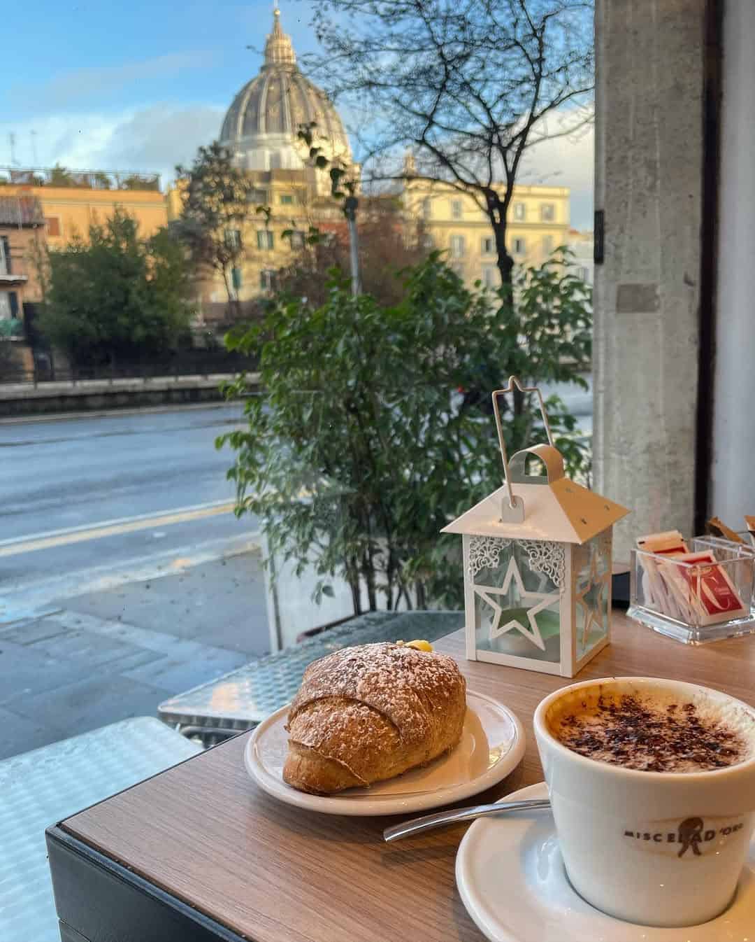 A cappuccino and croissant on a café table with a view of St. Peter’s Basilica outside the window—perfect for those exploring Gluten Free Restaurants in Rome.