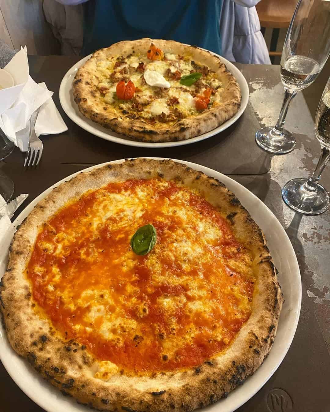 Two pizzas on white plates—one margherita and one with assorted toppings—served next to two glasses of sparkling wine, capturing the delicious experience at gluten free restaurants in Rome.