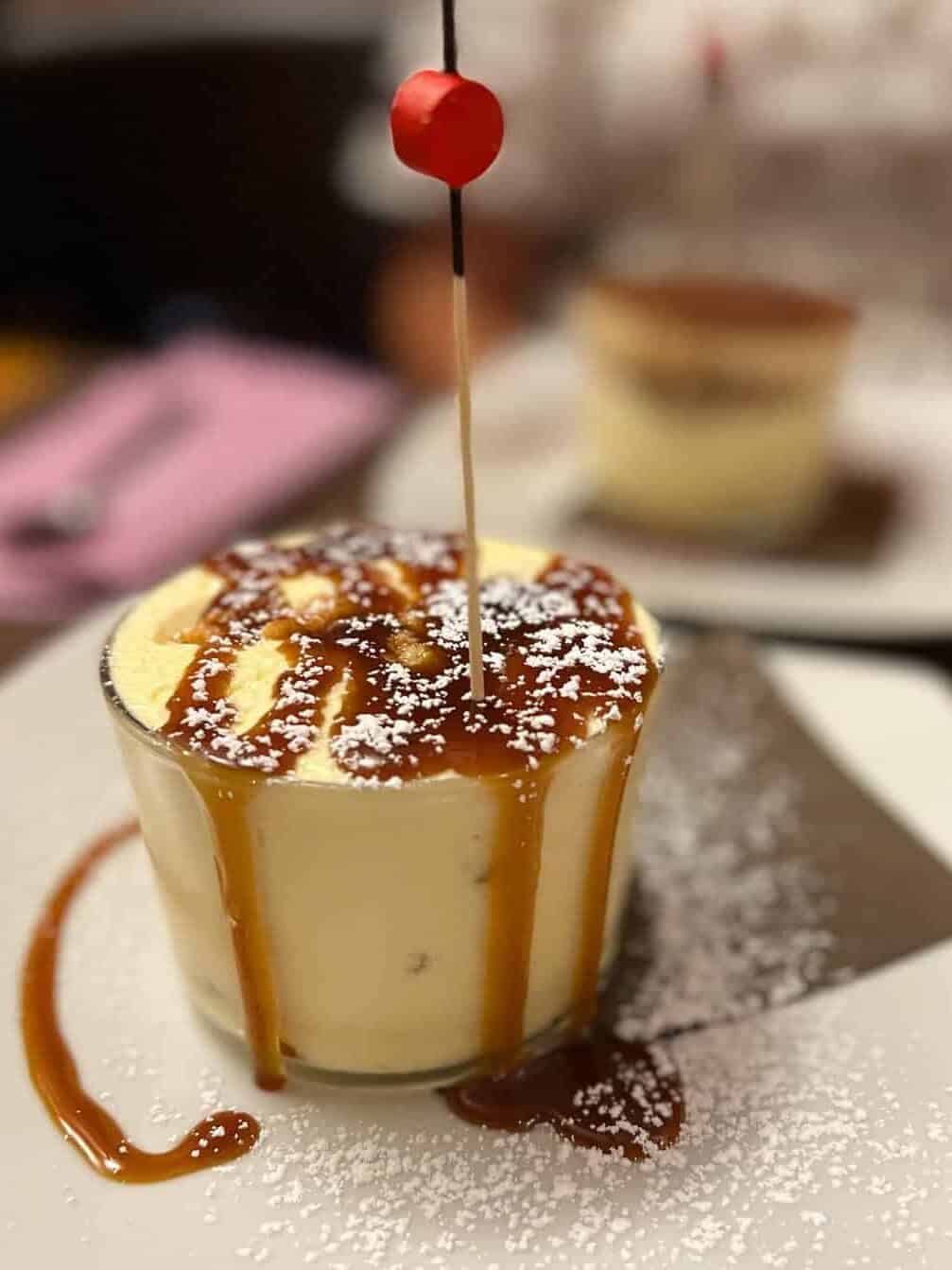 Glass of creamy dessert topped with caramel sauce, powdered sugar, and a red cherry on a stick, served on a white plate at one of the top Gluten Free Restaurants in Rome.