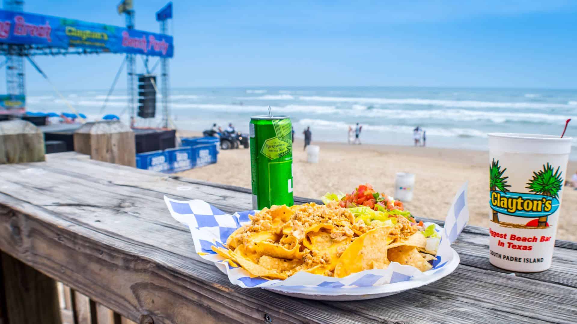 Plate of nachos, a drink can, and a cup on a wooden table overlooking a beach with people and a stage.
