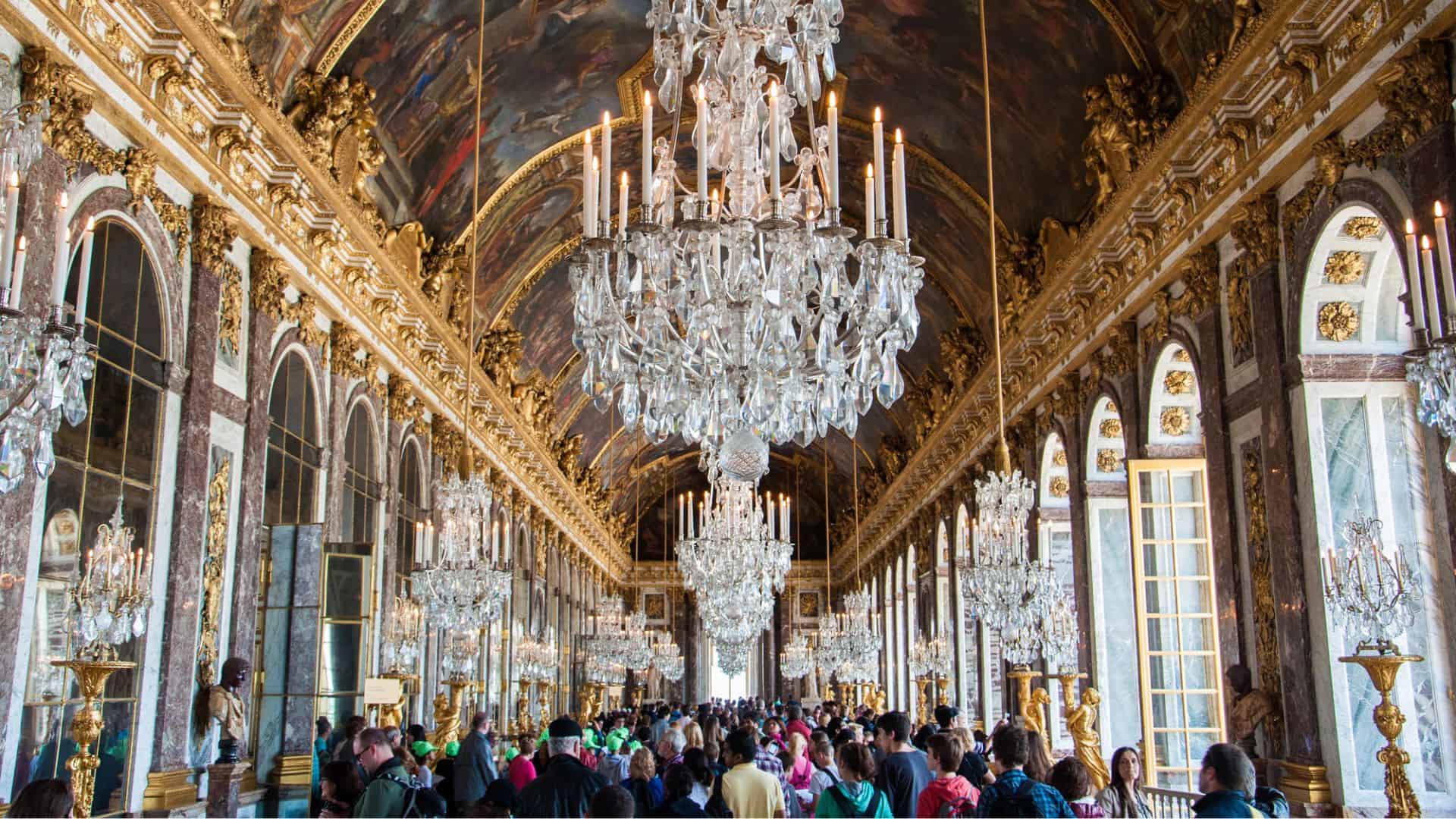 Crowds in the Hall of Mirrors at Versailles, with chandeliers, gilded decor, and painted vaulted ceiling.